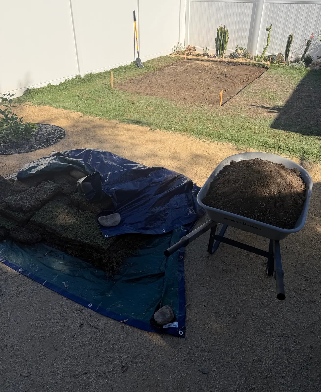 Stripped sod stacked on a tarp next to a wheelbarrow of soil, the cleared pad behind
